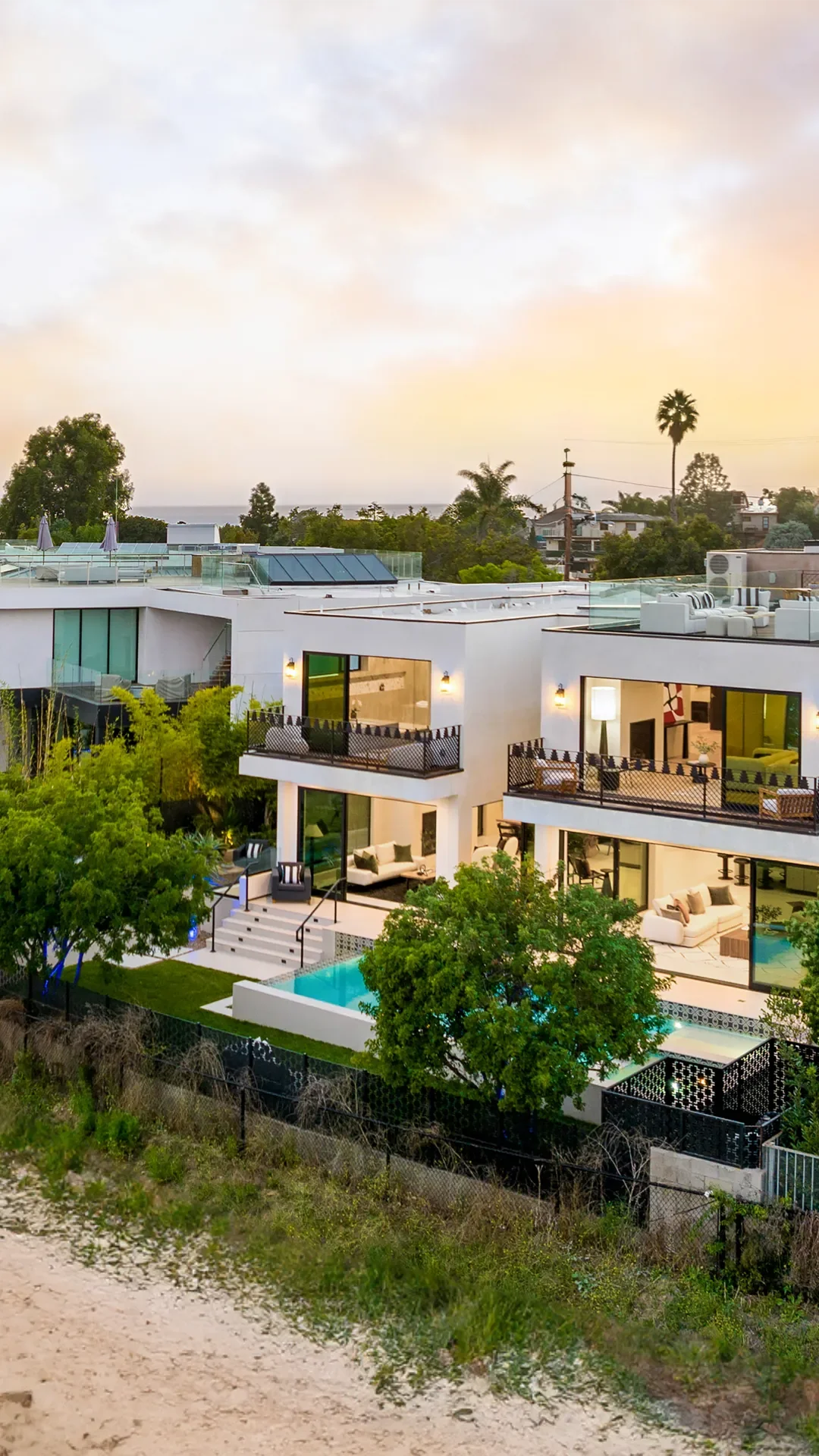 Modern white stucco beach home with pool, glass doors, metal railings, and rooftop terrace at dusk