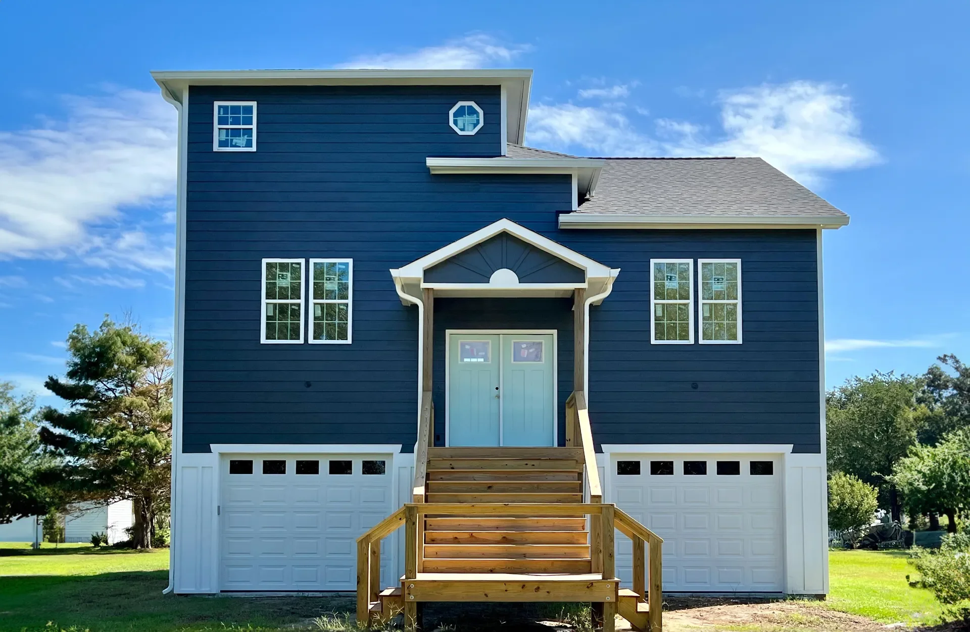 Dark blue siding home with white garage doors, wooden deck, and peaked entry portico