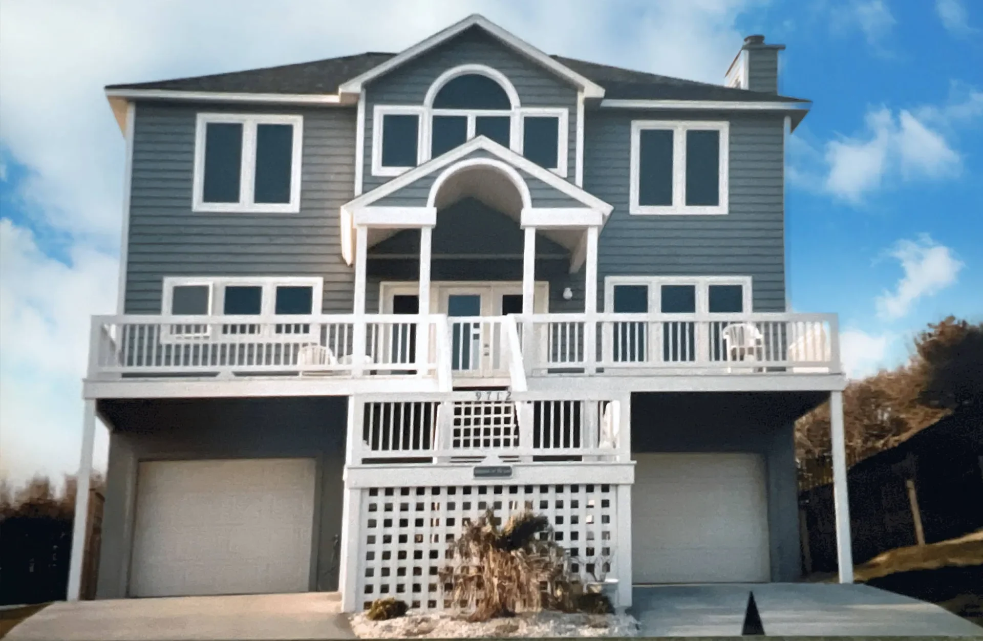 Gray siding elevated home with white deck railings, curved entry, and dual garages