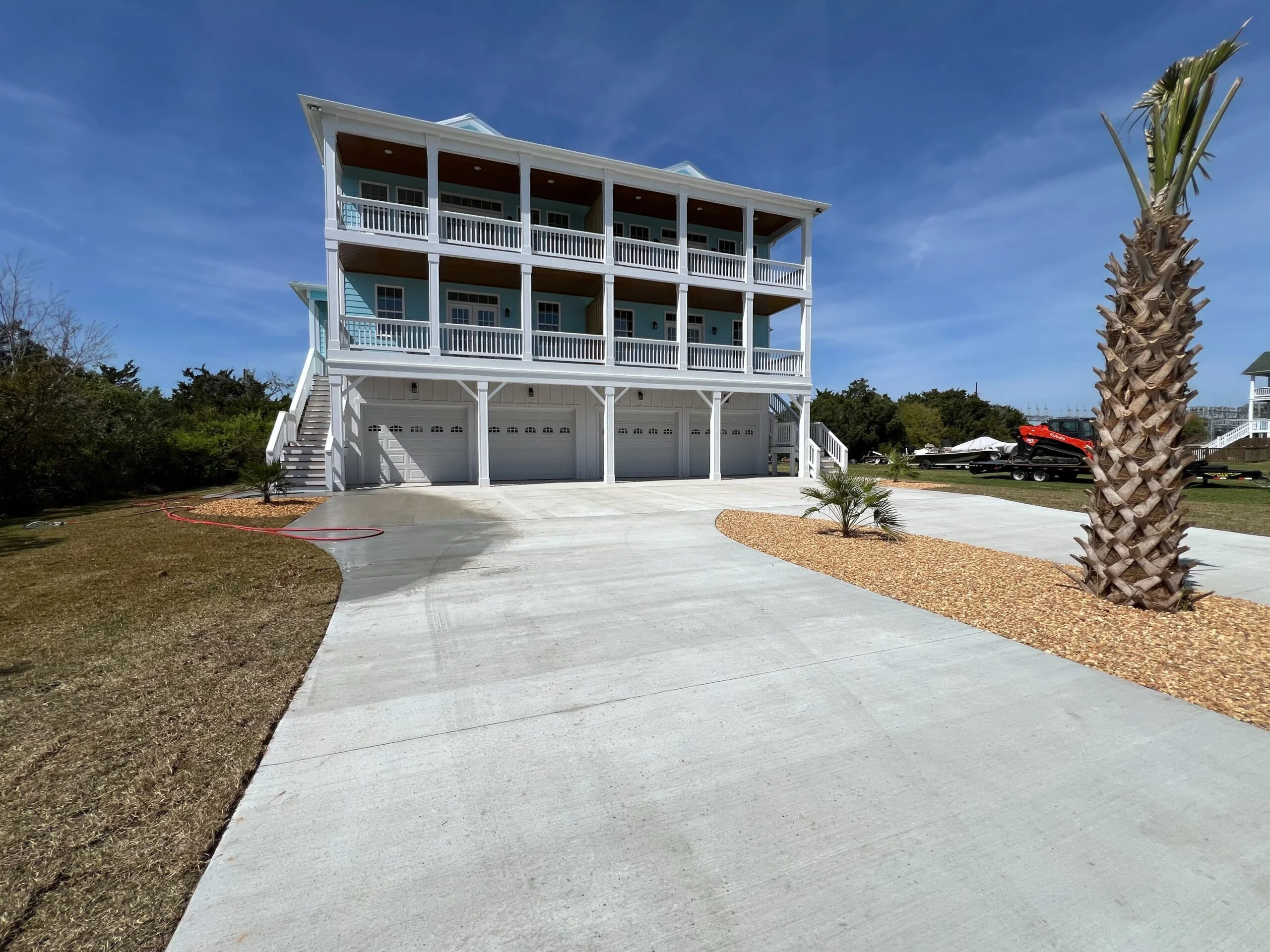 Three-story white elevated home with four-car garage, concrete driveway, and landscaped grounds